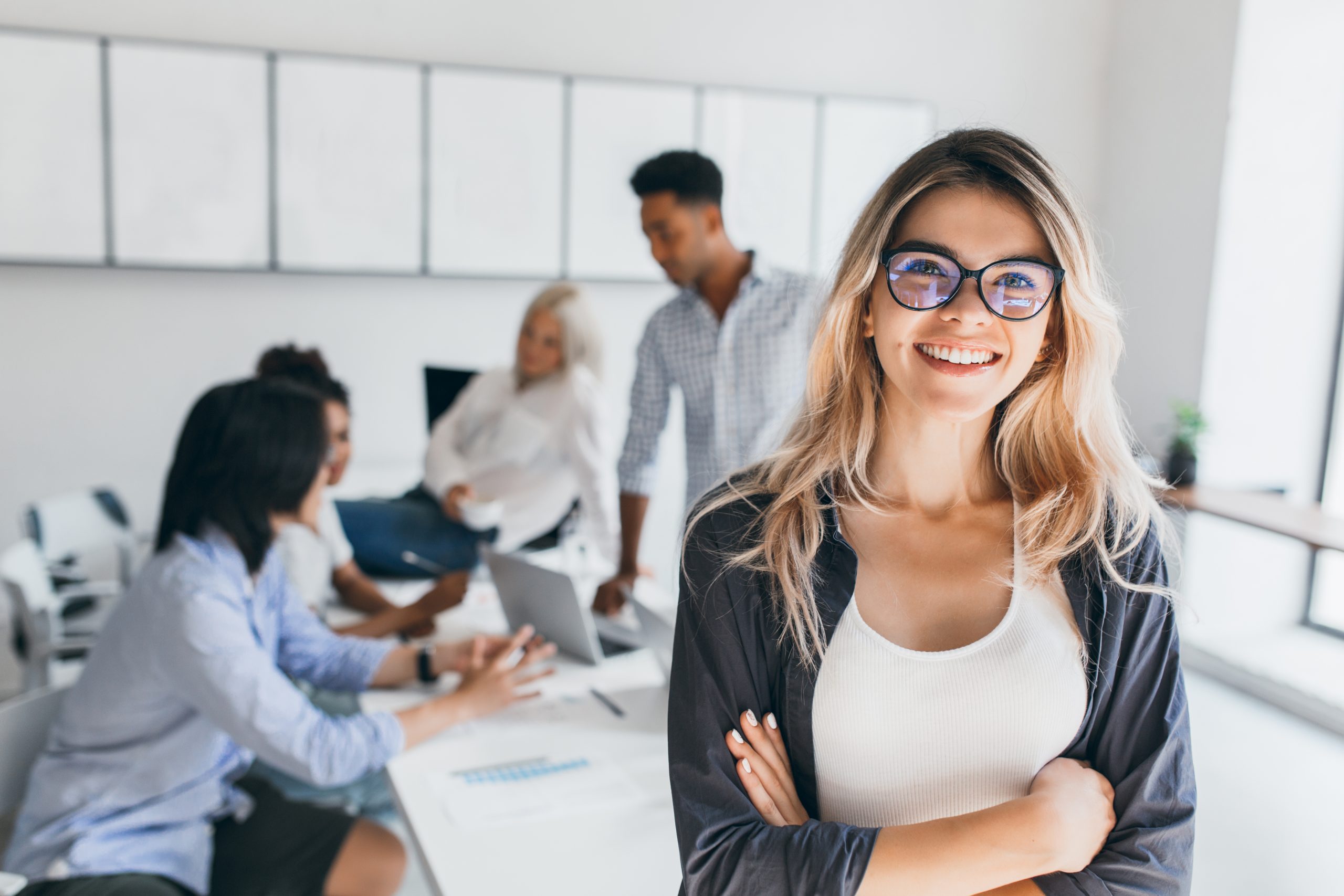 Blonde female executive posing with smile and arms crossed during brainstorm with managers. Indoor portrait of european student spending time in hall with asian and african friends..